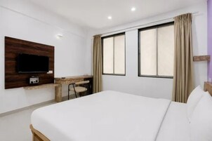 Habitación de hotel con cama, escritorio, silla, televisor y dos ventanas con cortinas.