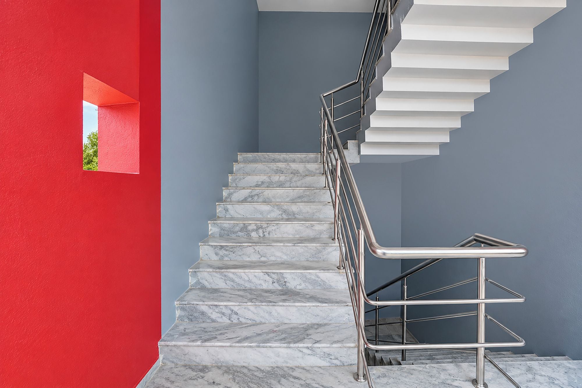 A staircase with marble steps and a metal handrail, leading to a landing with a red wall and a small window.