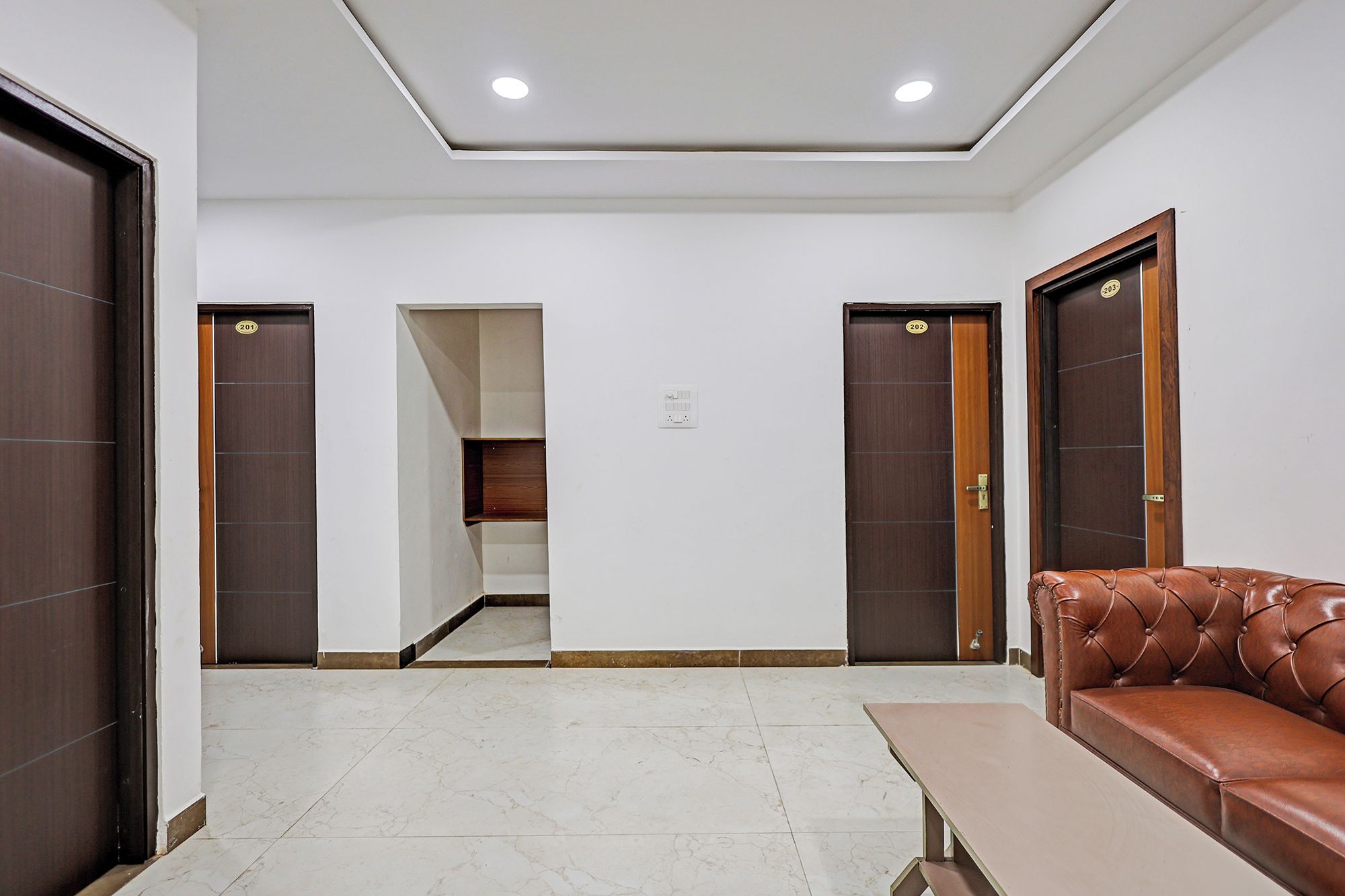 A hotel room with a brown leather sofa, a wooden door with a number, a white ceiling, and a tiled floor.