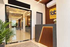 Zona de recepción de hotel con diseño moderno, que incluye un acceso por puerta de vidrio, un mostrador de recepción con panel de madera y un cartel con el nombre del hotel.