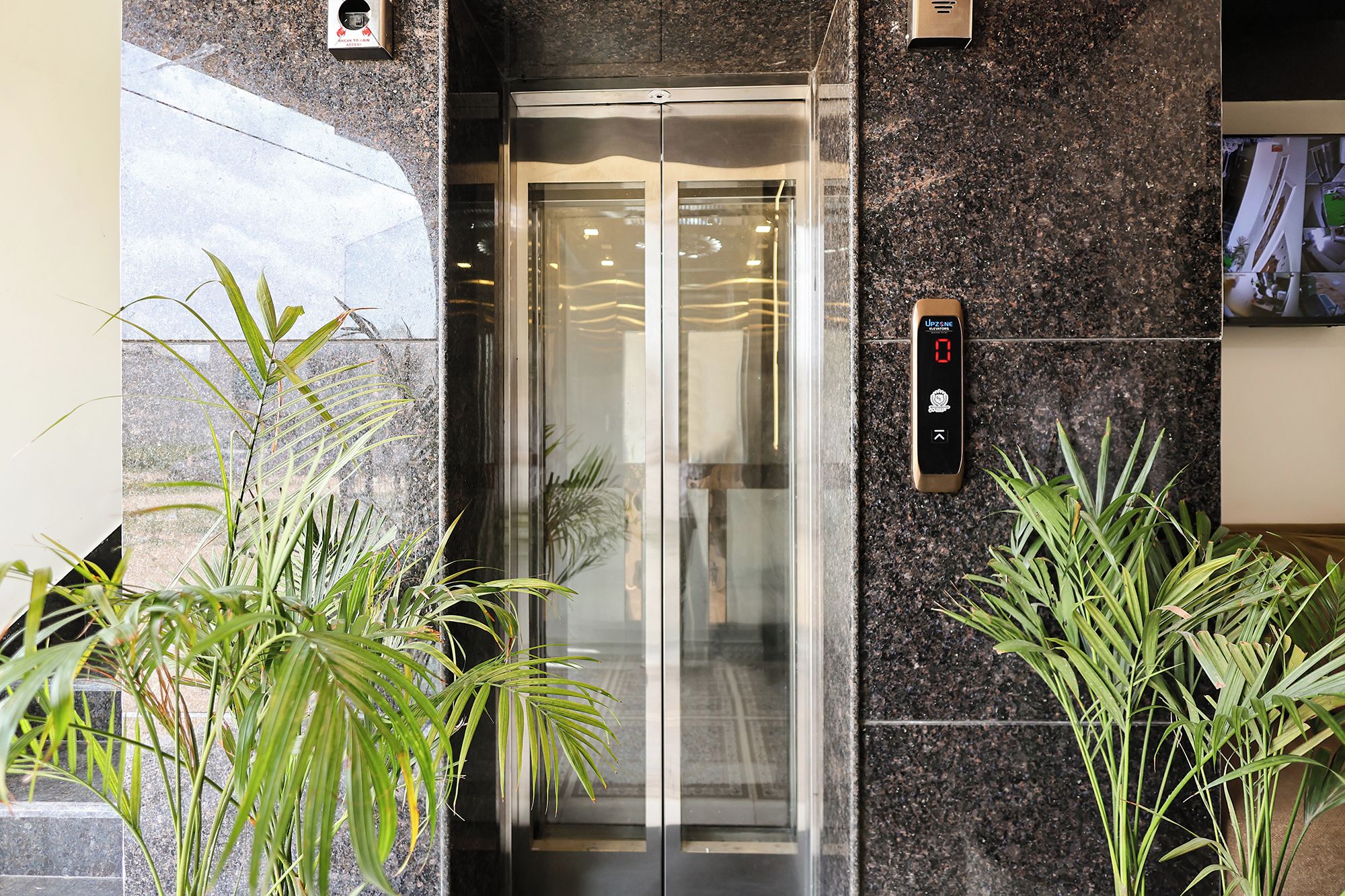 An elevator with a digital display, glass doors, and potted plants beside it.