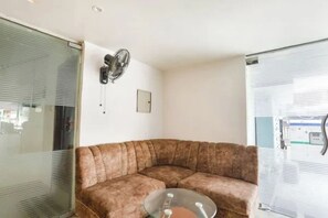 Een kamer met een hoekopstelling, een bruine bank, een ventilator aan de muur en een raam met zonwering.