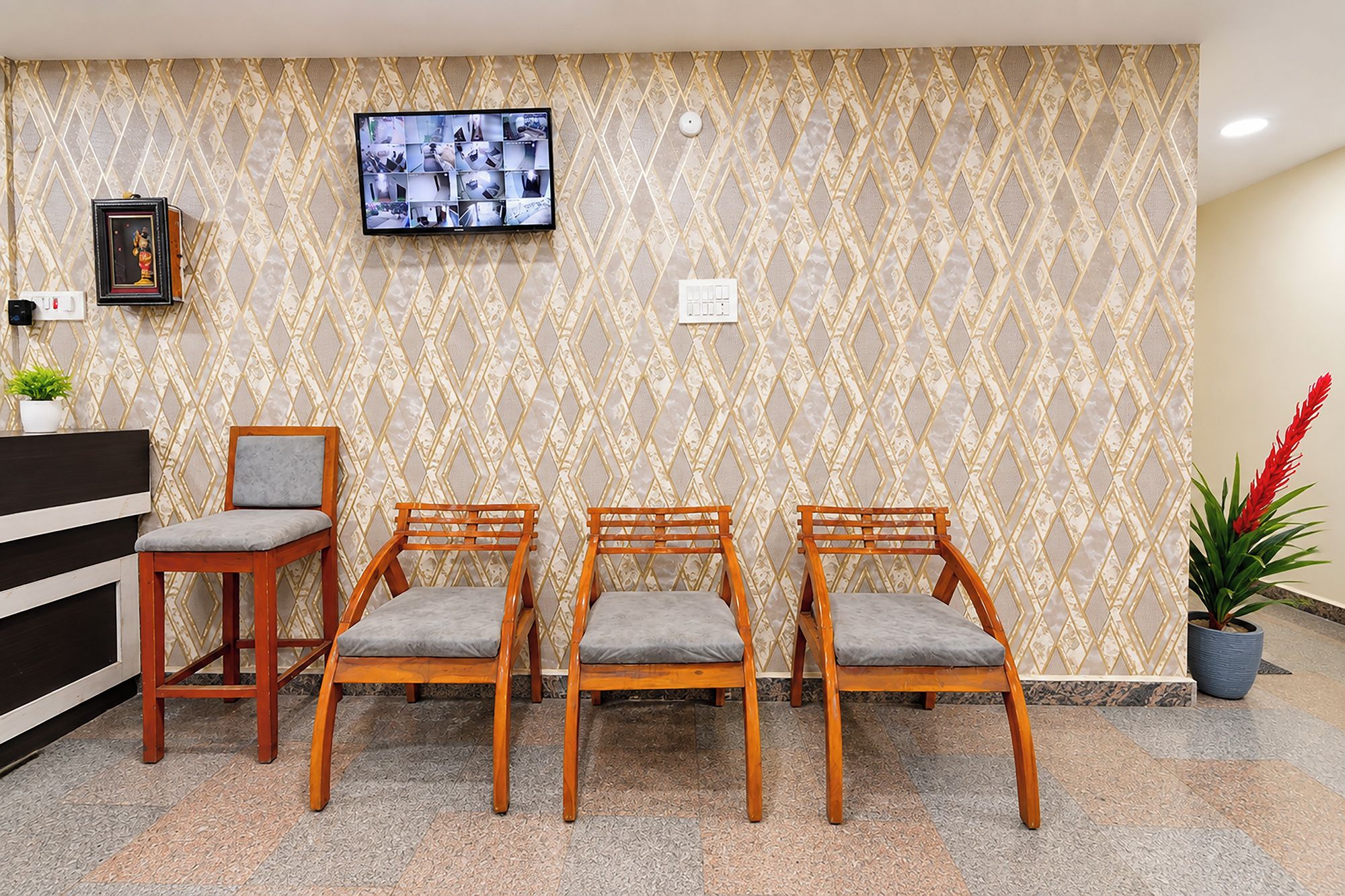 Una zona de recepción con un muro estampado, un televisor sobre un escritorio y tres sillas de madera con cojines grises.