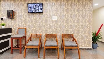 A reception area with a patterned wall, a television mounted above a desk, and three wooden chairs with grey cushions.