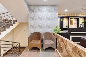 Un área de recepción con mostrador de mármol, dos sillas de mimbre, una escalera con barandillas de metal y una pared con un diseño geométrico.