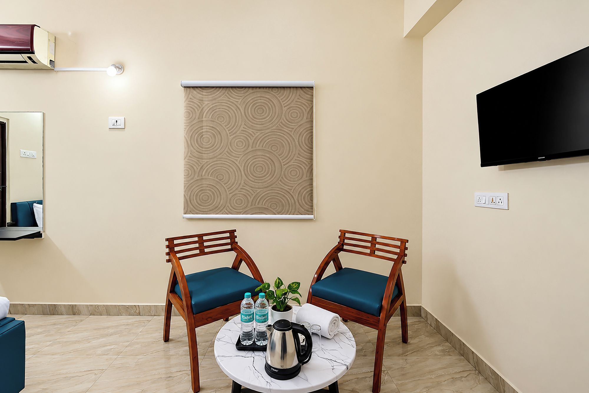 Una habitación con dos sillas de madera con cojines azules, una mesa redonda con una tetera, botellas de agua y una toalla enrollada. Un televisor está montado en la pared, y una persiana con diseño está sobre las sillas.