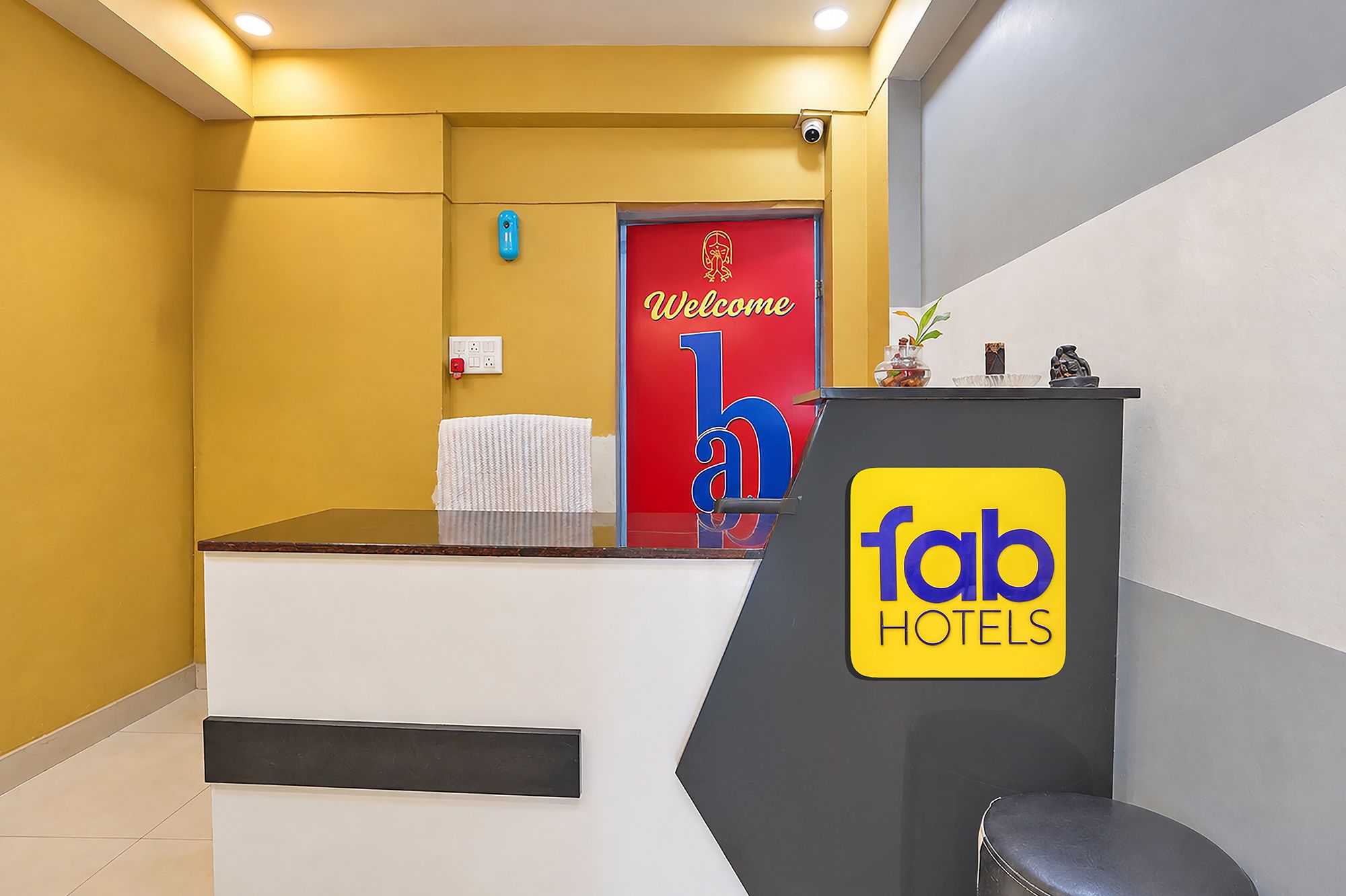 Una recepción con un cartel de 'fab hotels', una puerta roja con un letrero de bienvenida y una lámpara azul fijada a la pared.