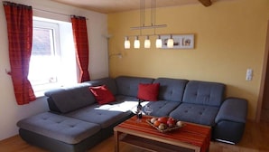 A living room with a grey sofa, red cushions, a wooden coffee table with fruit, and a window with curtains. - Holiday apartment in Vöhl with swimming pool (Vöhl)