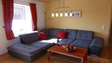 Ein Wohnzimmer mit grauem Sofa, roten Kissen, einem Holztisch mit Obst und einem Fenster mit Vorhängen.