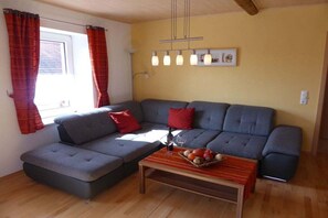 A living room with a grey sofa, red cushions, a wooden coffee table with fruit, and a window with curtains. - Holiday apartment in Vöhl with swimming pool (Vöhl)
