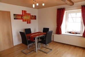 A dining area with a table and chairs, a radiator, a window with curtains, and a wall with a piece of abstract art. - Holiday apartment in Vöhl with swimming pool (Vöhl)