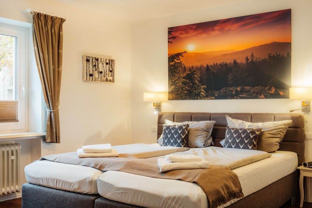 A bedroom with a bed, a window with curtains, a painting of a sunset, and a radiator.