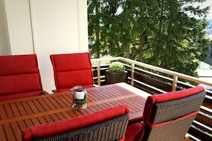 A wooden table with two red cushioned chairs, a glass pitcher with a green plant, and a view of trees and a building in the background. - Holiday apartment with 1 bedroom (Braunlage)