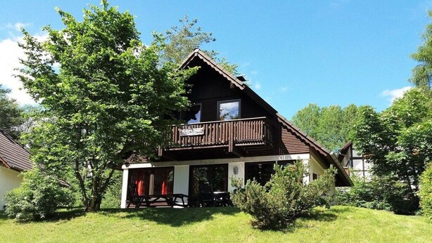 A two-story wooden house with a balcony, surrounded by greenery and trees. - Gingerbread House Gretel (Frankenau)
