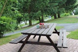 A wooden picnic table with a 'WELCOME' sign, surrounded by greenery and a paved path. - Gingerbread House Gretel (Frankenau)