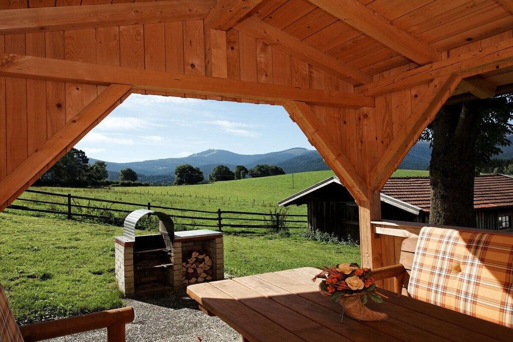 Un gazebo in legno con una panchina e un tavolo, affacciato su un prato con una montagna sullo sfondo.
