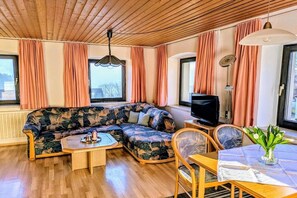 A living room with a sectional sofa, a television, a dining table with chairs, and a window with curtains. - Holiday apartment with 1 bedroom (Lohberg)