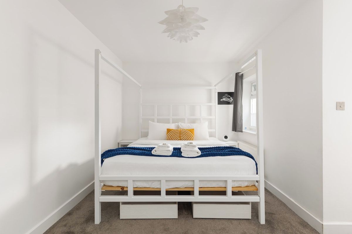 Una cama con dosel blanco, con una manta azul y dos toallas, un ventilador de techo blanco y una ventana pequeña con cortina.