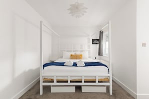 A white four-poster bed with a blue blanket and two towels, a white ceiling fan, and a small window with a curtain. - New! Modern 2BR Flat | Forest Gate (Greater London)