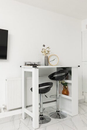 A white bar stool with a black seat and a metal base, a small round table with a clock, and a vase with a plant on a white shelf. - New! Spacious 4BR House w/Garden in Mitcham (Greater London)