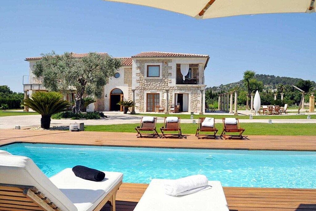 Un espace aménagé au bord de la piscine, avec des chaises longues et une vue dégagée sur un bâtiment à deux étages, doté d’une façade en pierre et d’un toit recouvert de tuiles.
