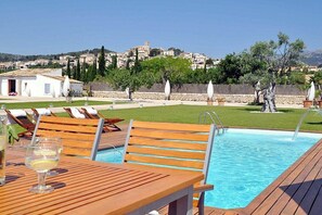 A pool area with wooden loungers and a table, a clear view of a hillside town, and a stone wall with trees. - Ses Rentedores [2257] (Selva)