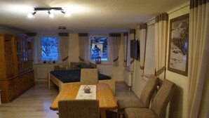 Una habitación con una mesa de comedor, sillas, un sofá, un televisor y ventanas con cortinas.