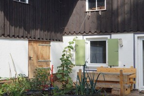 Una porta de fusta, una finestra amb persiana verda i una banca de fusta en un jardí.