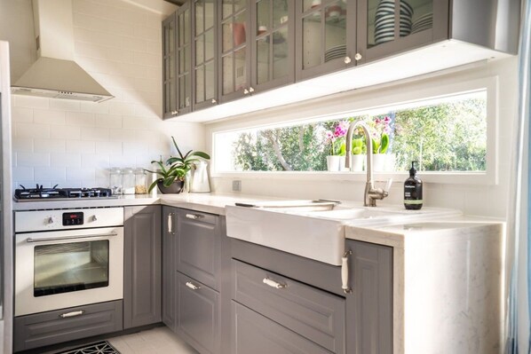 A modern kitchen with a gas stove, oven, grey cabinets, a window with a view of greenery, and a sink with a faucet.