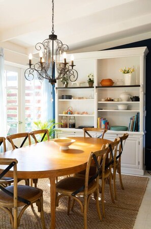 A dining area with a round wooden table, six chairs, a chandelier, and a white cabinet with shelves. - Chiara`s Art Cottage 1BR Coastal Garden Retreat (Sorrento)