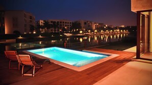 An outdoor swimming pool with wooden deck and lounge chairs, surrounded by buildings and a body of water reflecting lights. - Private Pool Villa in Hawana Salalah — 5 min to the Beach (Taqah)