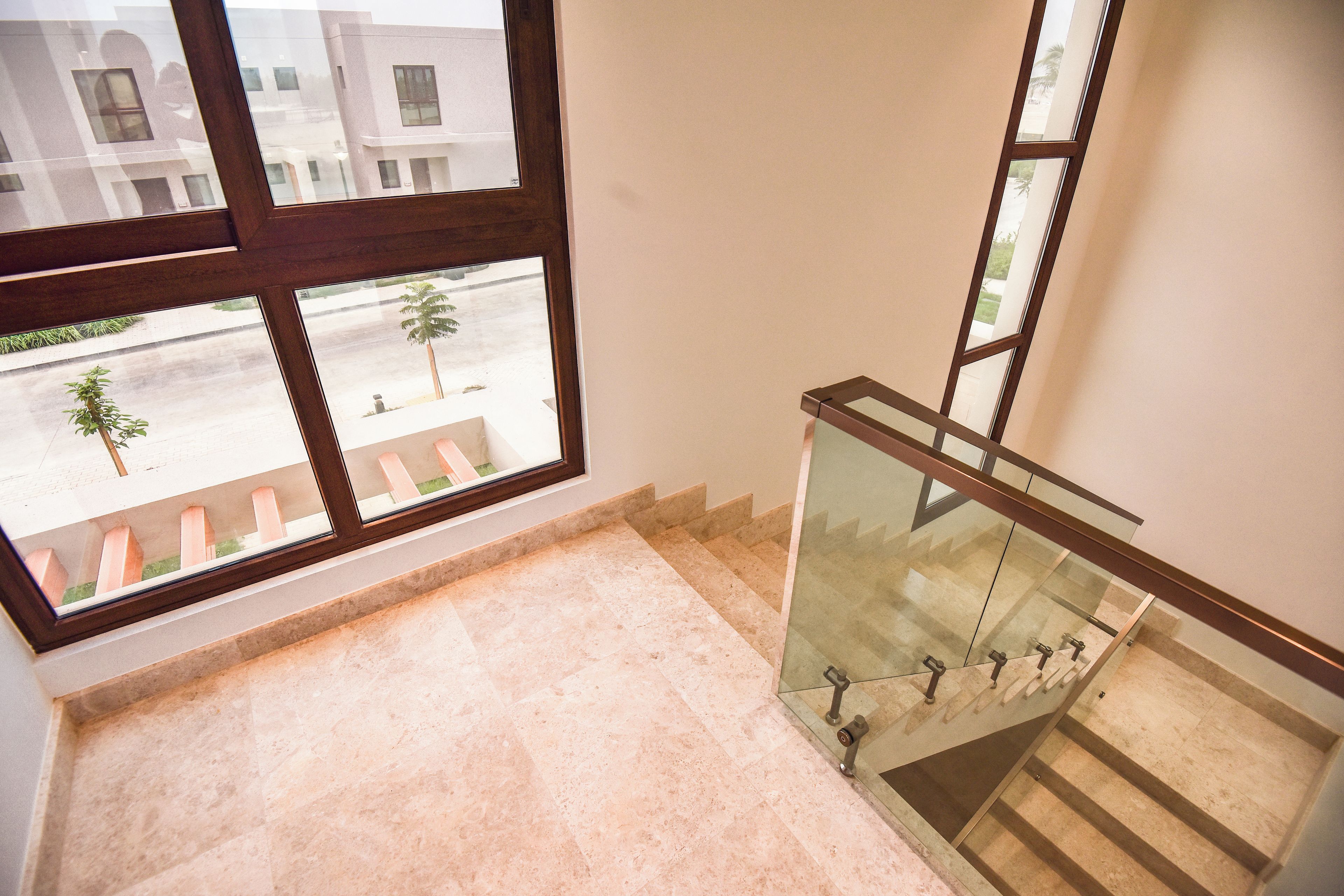 A staircase with glass railings leading to an upper floor, a view of a street with palm trees, and a building reflection in the window.