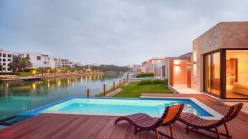 A swimming pool with lounge chairs, a river view, and modern buildings in the background.