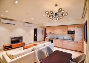 A modern living room with a flat-screen TV, sofa, and dining table. A kitchen with wooden cabinets and stainless steel appliances is visible. - Private Pool Villa in Hawana Salalah — 5 min to the Beach (Taqah)