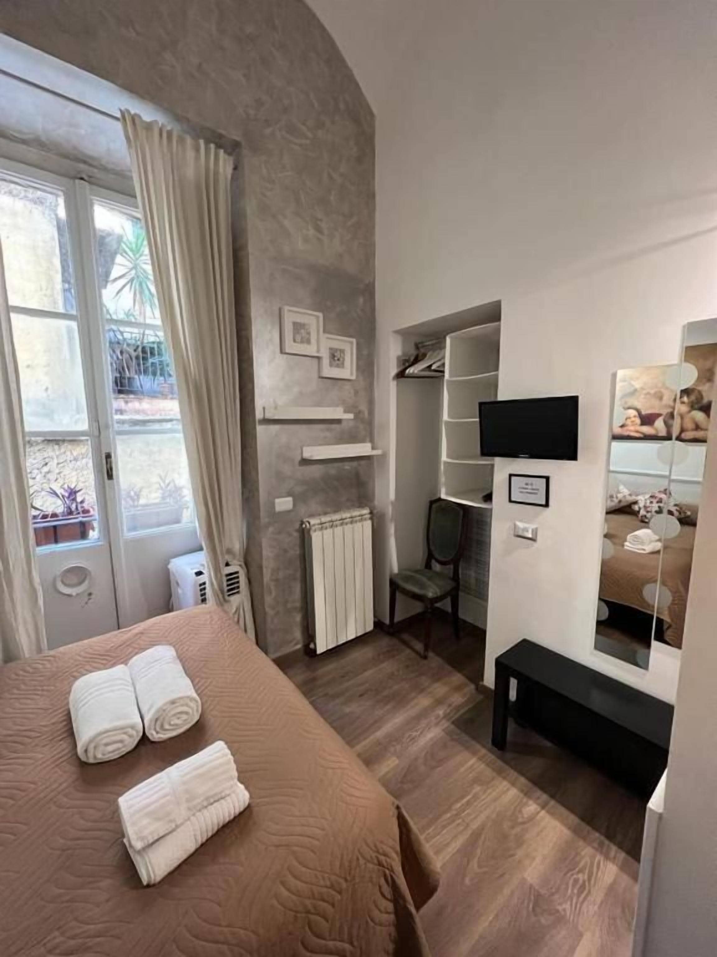 Une chambre d’hôtel avec un lit, une chaise, une télévision, un miroir et une fenêtre avec des rideaux.