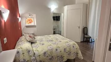 Ein Schlafzimmer mit einem Bett, einem Schreibtisch und einem Sessel. Eine Tür und ein Spiegel sind an der Wand angebracht.