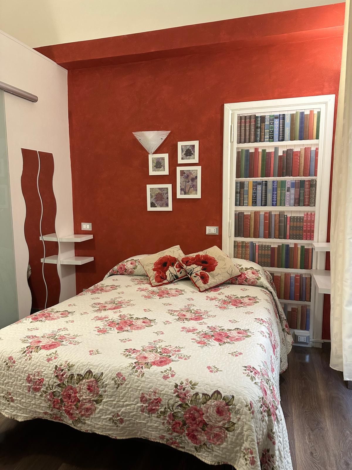 A bedroom with a red accent wall, a bookshelf, and a bed with floral bedding.
