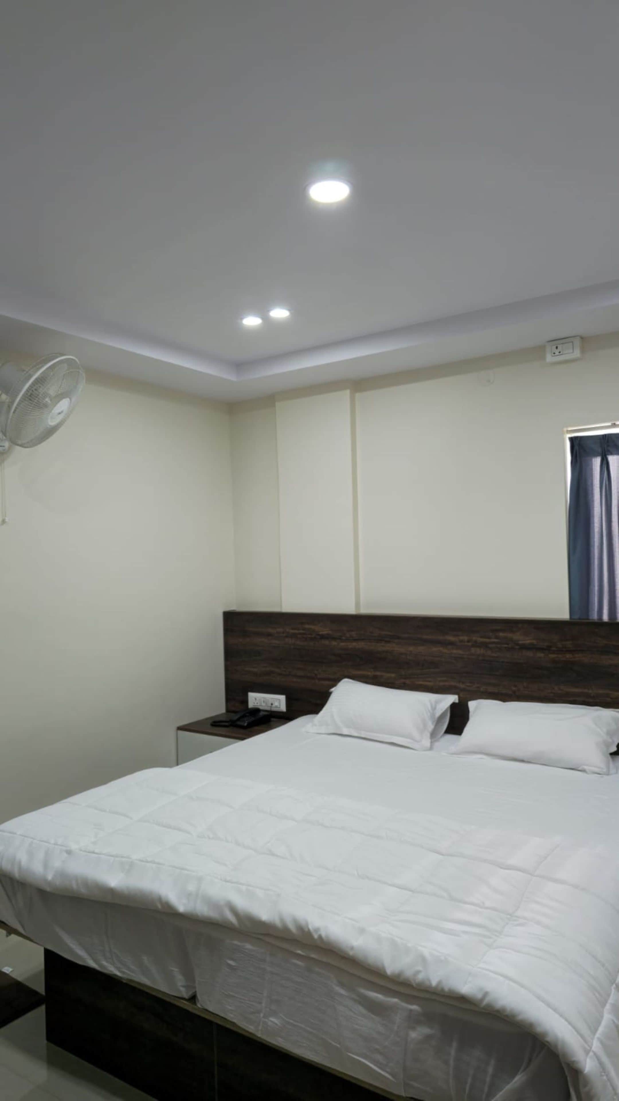 A bedroom with a bed, a nightstand, a wall-mounted fan, and recessed ceiling lighting.