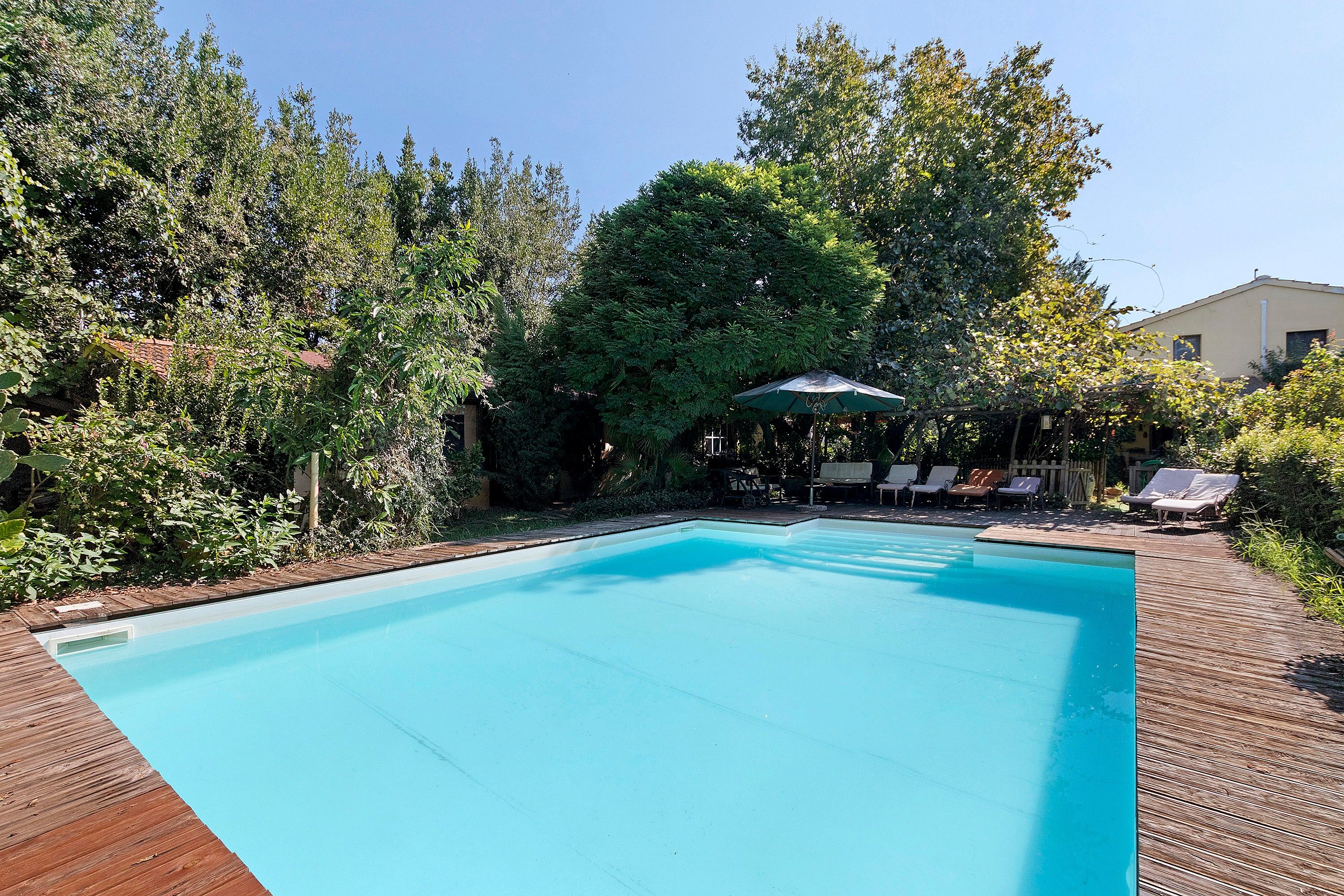 A rectangular swimming pool with a wooden deck surrounding it. There are lounge chairs and a table with an umbrella in the background.