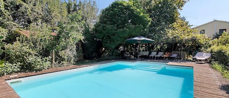 Une piscine rectangulaire entourée d’une terrasse en bois. On aperçoit, au fond, des chaises longues et une table avec un parasol.