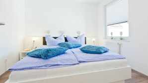 A bedroom with a white bed, blue pillows, a nightstand with a lamp, and a window with a view of the outside. - Holiday apartment with 2 bedrooms (Norderney)