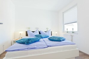 Ein Schlafzimmer mit einem weißen Bett, blauen Kissen, einem Nachttisch mit Lampe und einem Fenster mit Blick nach draußen.