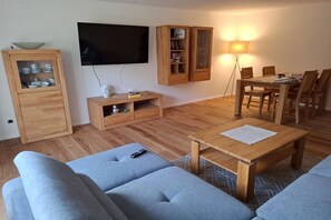 Ett vardagsrum med en soffa, ett soffbord, en tv som är monterad på väggen, träskåp och ett matbord med stolar.
