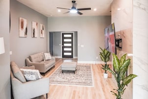 Una sala de estar moderna con sofá, sillón y otoman, ventilador de techo y un ventanal con persianas.