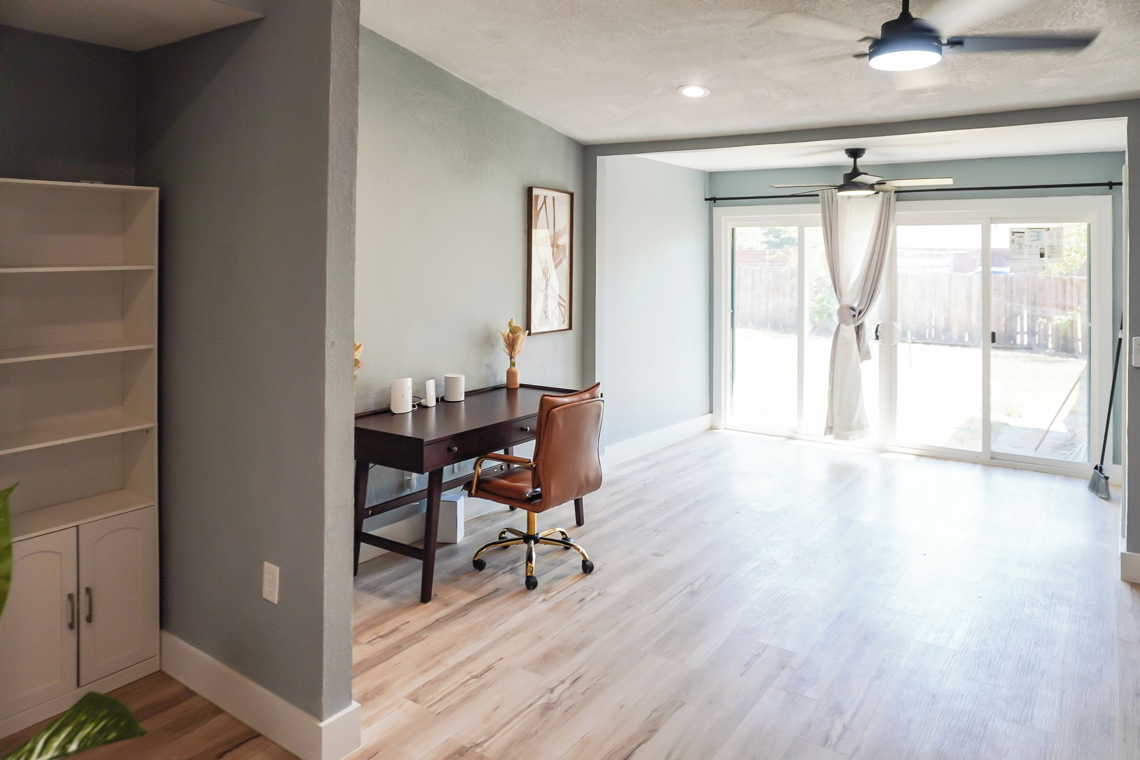 A spacious room with a wooden floor, a desk with a chair, a ceiling fan, and a large sliding glass door leading to an outdoor area.