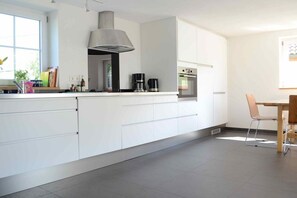 A modern kitchen with white cabinetry, a stainless steel range hood, and a dining area with a wooden table and chairs. - Black Forest Lounge Holiday Home Lindenbuch (Loßburg)