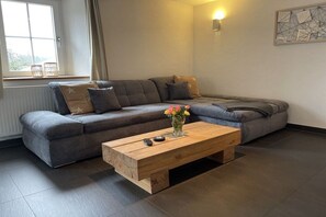 A modern living room with a grey sectional sofa, a wooden coffee table with a vase of flowers, and a wall-mounted light fixture. - Black Forest Lounge Holiday Home Lindenbuch (Loßburg)