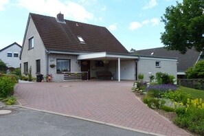 A two-story house with a covered porch, a paved driveway, and a garden with various plants and flowers. - Holiday apartment with 1 bedroom (Heiligenhafen)