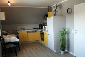 Een kleine keuken met een eettafel, gele kastjes, een koelkast en een potplant.
