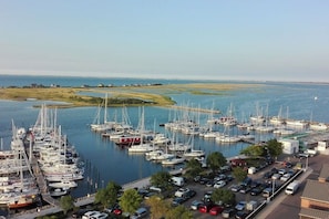 A marina with multiple boats docked, a parking area with cars, and a view of the water and distant shoreline. - Holiday apartment with 1 bedroom (Heiligenhafen)